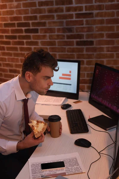 Hombre de negocios con pizza y café para llevar mirando la computadora cerca del periódico y el teléfono inteligente por la noche - foto de stock