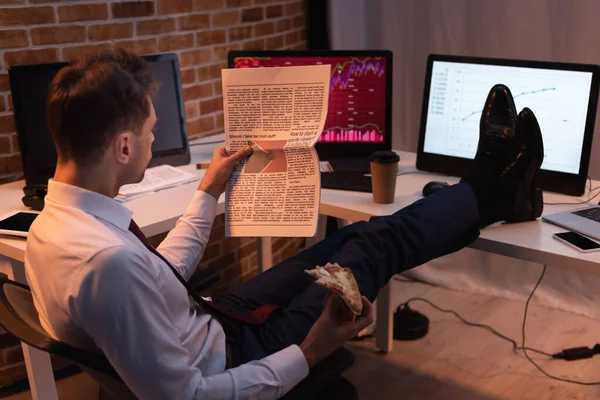 Uomo d'affari che legge giornale e tiene pizza da asporto vicino a computer in sera — Foto stock
