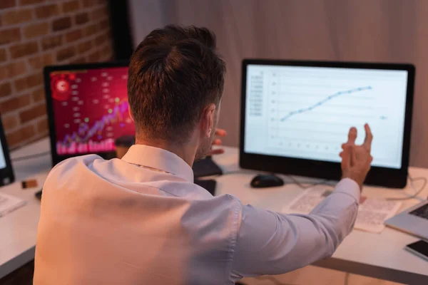 Обратный вид бизнесмена, указывающего на компьютер с финансовыми диаграммами на размытом фоне — стоковое фото