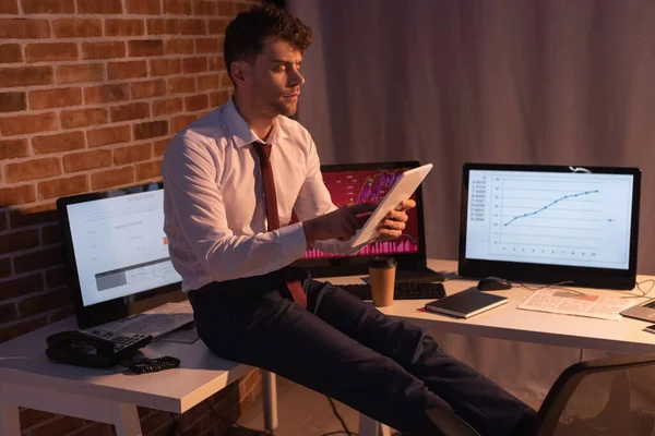 Uomo d'affari che utilizza tablet digitale vicino a computer, telefono e giornale su sfondo sfocato in serata — Foto stock