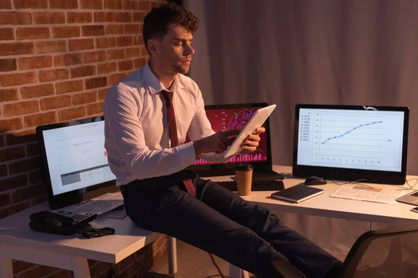 Geschäftsmann mit digitalem Tablet in der Nähe von Computern, Telefon und Zeitung auf verschwommenem Hintergrund am Abend — Stockfoto