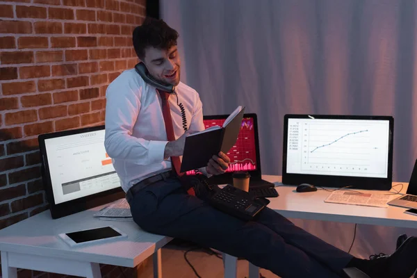 Hombre de negocios sonriente hablando por teléfono y sosteniendo un cuaderno cerca de dispositivos en la oficina - foto de stock