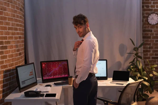 Businessman adjusting tie near computers and newspaper in office in evening — Stock Photo