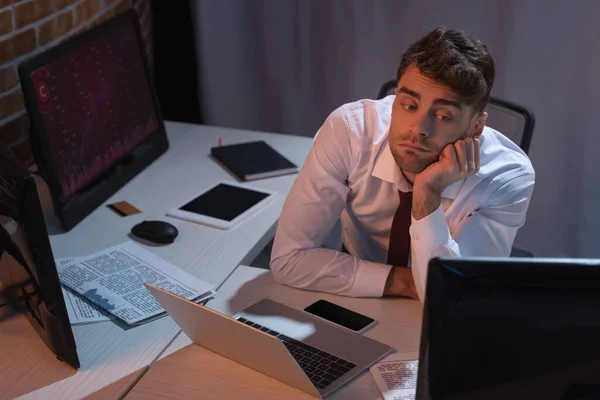 Empresário olhando para o computador perto de dispositivos e jornais à noite — Fotografia de Stock