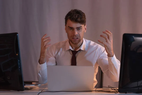 Hombre de negocios agresivo mirando a la cámara cerca de computadoras en primer plano borroso - foto de stock