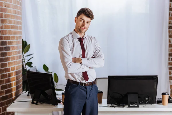 Uomo d'affari in piedi con le braccia incrociate vicino ai computer in ufficio — Foto stock