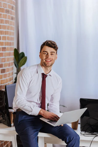 Allegro uomo d'affari che utilizza laptop vicino al caffè da asporto e computer in ufficio — Stock Photo