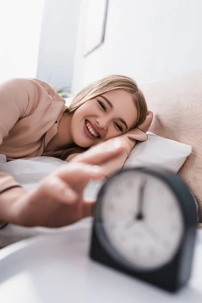 Happy young woman reaching alarm clock on bedside table and blurred foreground — Stock Photo