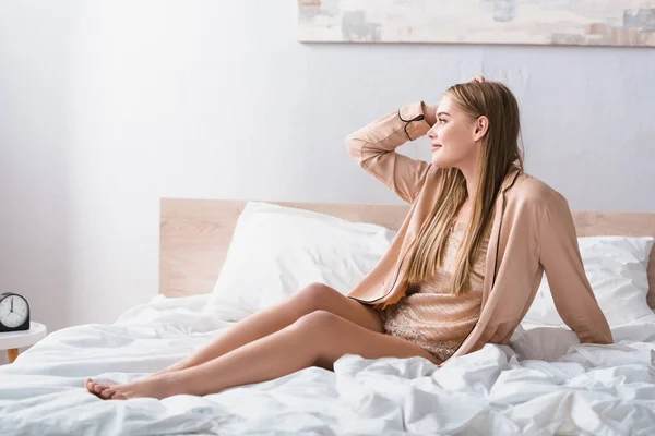 Freudige junge Frau schaut weg, während sie im modernen Schlafzimmer auf dem Bett sitzt — Stockfoto