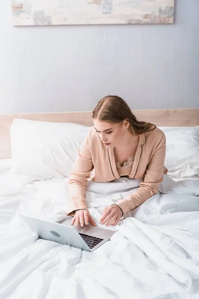 Joven freelancer en bata de seda usando portátil en el dormitorio - foto de stock