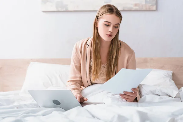 Joven freelancer en bata de seda mirando documentos cerca del portátil en la cama - foto de stock