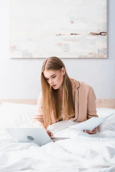 Young freelancer in silk robe using laptop and holding papers in bedroom — Stock Photo
