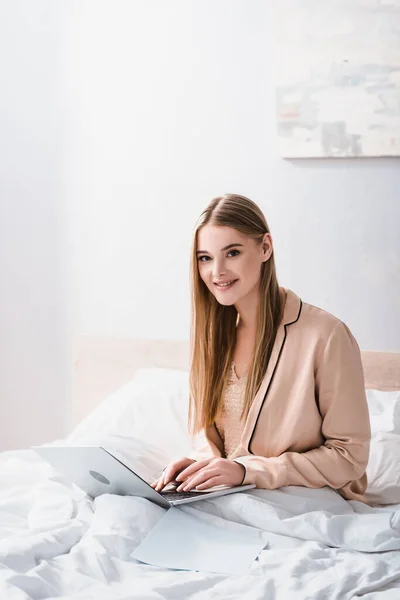 Alegre joven freelancer en bata de seda utilizando el ordenador portátil en el dormitorio - foto de stock