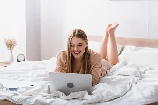 Smiling young freelancer in silk robe lying on bed and using laptop — Stock Photo