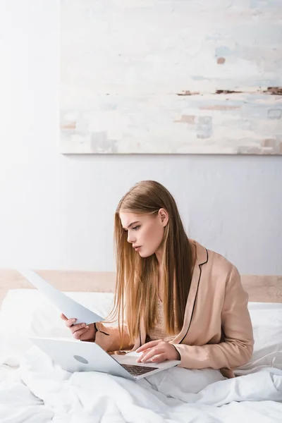 Young woman in silk robe looking at document neat laptop on bed — Stock Photo