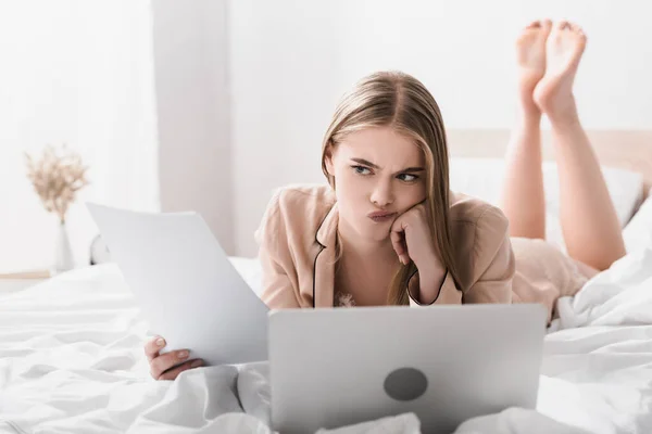 Mujer disgustada en bata de seda acostada en la cama, mirando el papel cerca de la computadora portátil en primer plano borrosa - foto de stock