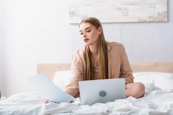 Joven freelancer en bata de satén mirando el documento cerca de la computadora portátil en el dormitorio - foto de stock