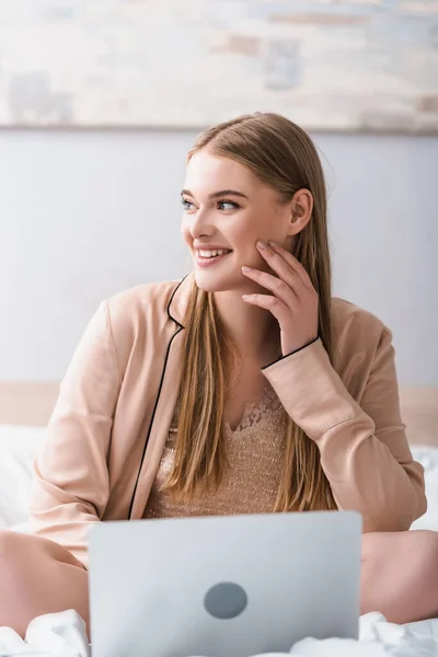 Smiling freelancer in satin robe looking away near laptop in bedroom — Stock Photo