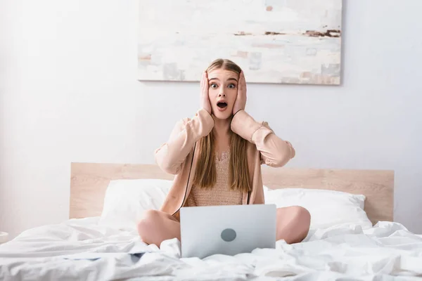 Shocked freelancer in satin robe looking at camera near laptop in bedroom — Stock Photo