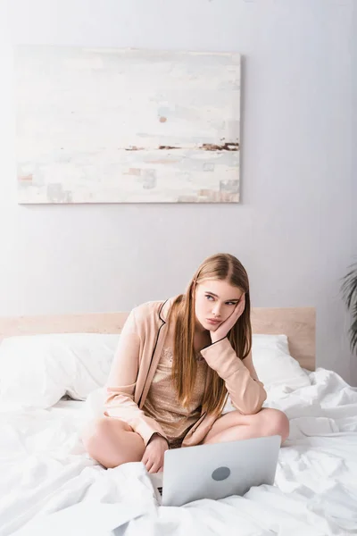 Mujer joven aburrida en bata de satén mirando lejos cerca de la computadora portátil en la cama - foto de stock