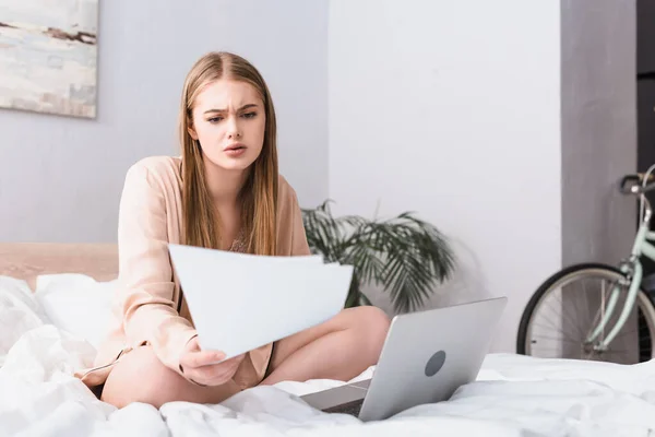 Joven freelancer en bata de seda mirando documentos cerca del portátil en la cama - foto de stock