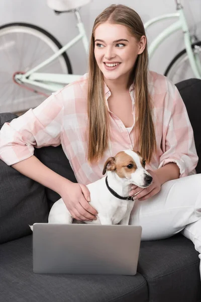 Femme heureuse caressant jack russell terrier près de l'ordinateur portable sur le canapé — Photo de stock