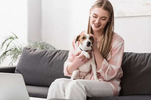 Sorrindo mulher segurando em braços jack russell terrier enquanto sentado no sofá com laptop — Fotografia de Stock