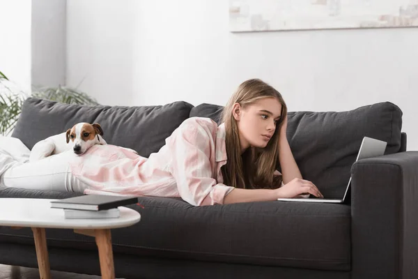 Giovane donna sdraiata sul divano con jack russell terrier e utilizzando il computer portatile in soggiorno — Foto stock