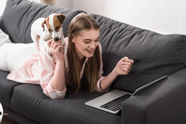 Femme heureuse couché sur le canapé avec jack russell terrier et en utilisant un ordinateur portable dans le salon — Photo de stock