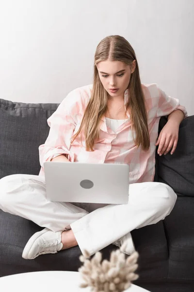 Joven freelancer sentado con las piernas cruzadas en el sofá y utilizando el ordenador portátil - foto de stock