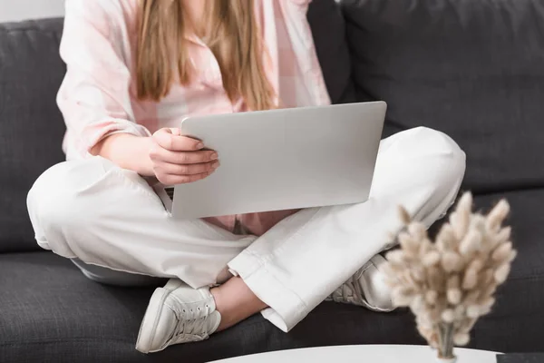 Cropped view of young freelancer sitting with crossed legs on couch and using laptop — Stock Photo