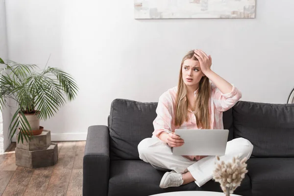 Besorgter junger Freiberufler sitzt mit gekreuzten Beinen auf Couch und benutzt Laptop — Stockfoto