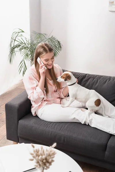 Happy woman sitting on couch with jack russell terrier and talking on smartphone — Stock Photo