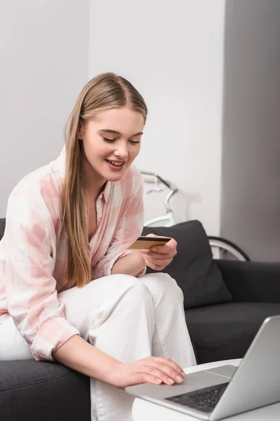 Jeune femme heureuse tenant la carte de crédit et utilisant un ordinateur portable sur la table basse — Photo de stock