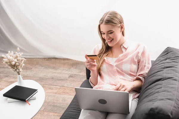 Happy young woman holding credit card near laptop while sitting on sofa — Stock Photo