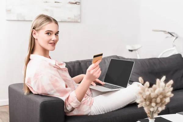 Feliz mujer joven sosteniendo la tarjeta de crédito cerca del ordenador portátil con pantalla en blanco - foto de stock