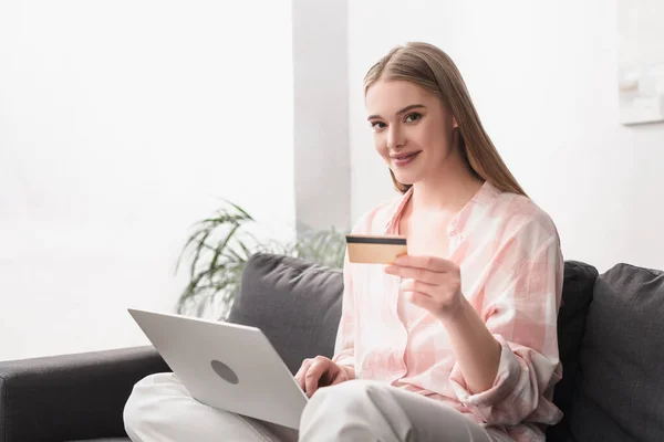 Молодая улыбающаяся женщина держит кредитную карту возле ноутбука во время покупок в Интернете дома — стоковое фото