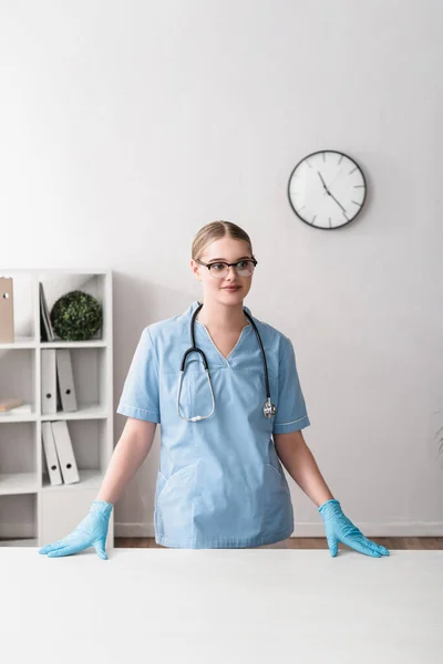 Happy young veterinarian in glasses and latex gloves standing near workplace — Stock Photo