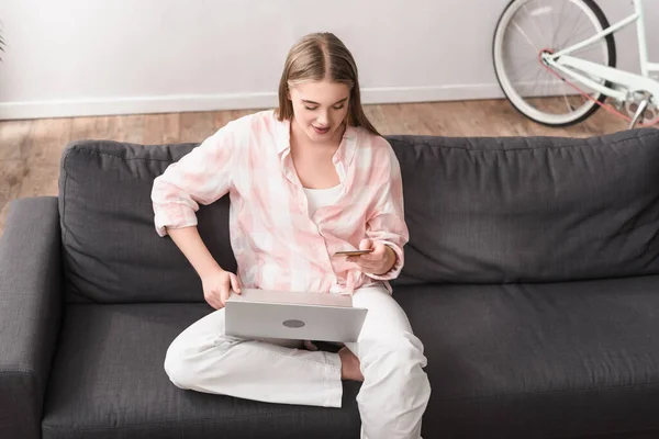 Mujer que sostiene la tarjeta de crédito cerca del ordenador portátil mientras que e-compras en casa - foto de stock