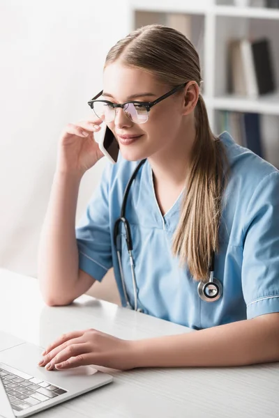 Veterinário sorrindo em óculos falando no smartphone perto de laptop na mesa — Fotografia de Stock