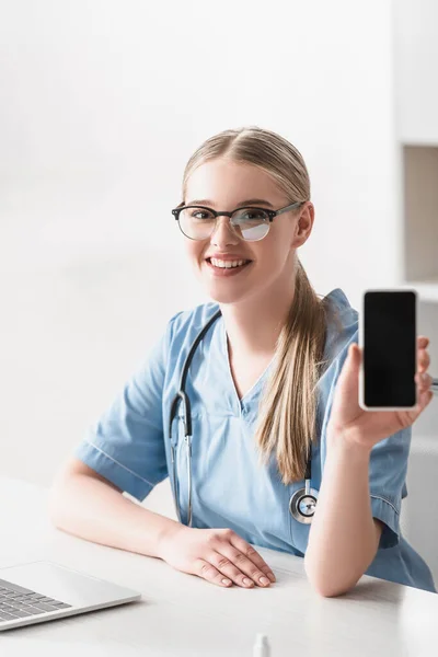 Cheerful veterinarian in glasses holding smartphone with blank screen near laptop on desk — Stock Photo