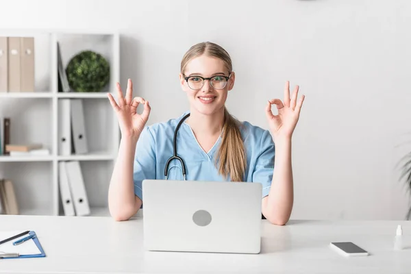 Cheerful veterinarian in glasses showing ok sign near gadgets on desk — Stock Photo