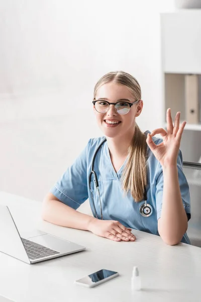 Cheerful veterinarian in glasses showing ok sign near gadgets on table — Stock Photo