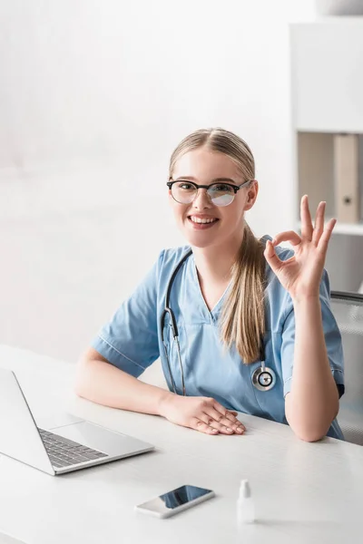 Cheerful veterinarian in glasses showing ok sign near gadgets on table — Stock Photo