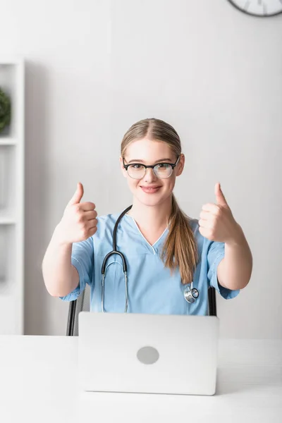 Veterinario alegre en gafas que muestran los pulgares hacia arriba cerca de la computadora portátil en el escritorio - foto de stock