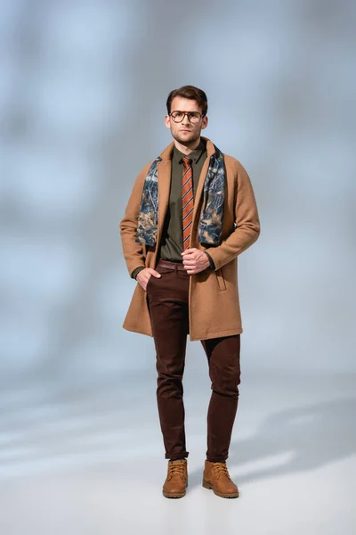 Повна довжина модного чоловіка в зимовому пальто, що стоїть з рукою в кишені на сірому — стокове фото