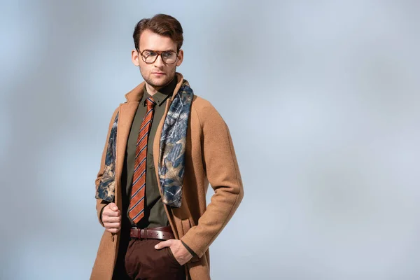 Стильный мужчина в очках и зимнем пальто, стоящий с рукой в кармане на сером — стоковое фото