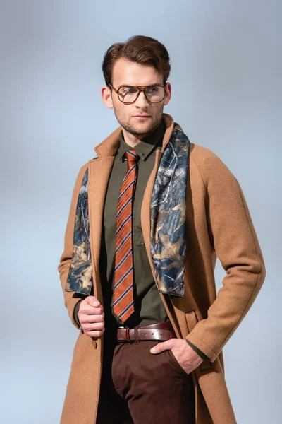Hombre de moda en gafas y abrigo de invierno de pie con la mano en el bolsillo en gris - foto de stock