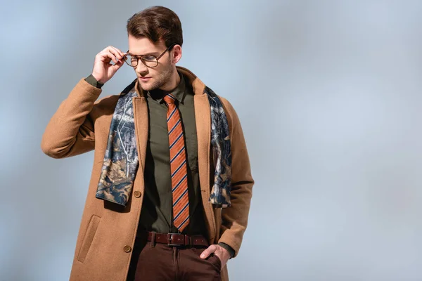 Hombre de moda ajustando gafas y de pie en abrigo de invierno con la mano en el bolsillo en gris - foto de stock