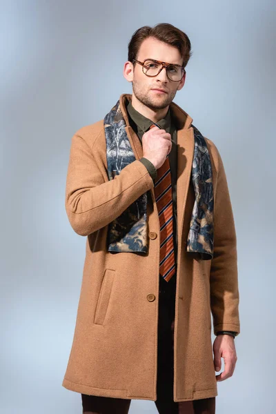 Trendy man adjusting tie and standing in winter coat on grey — Stock Photo